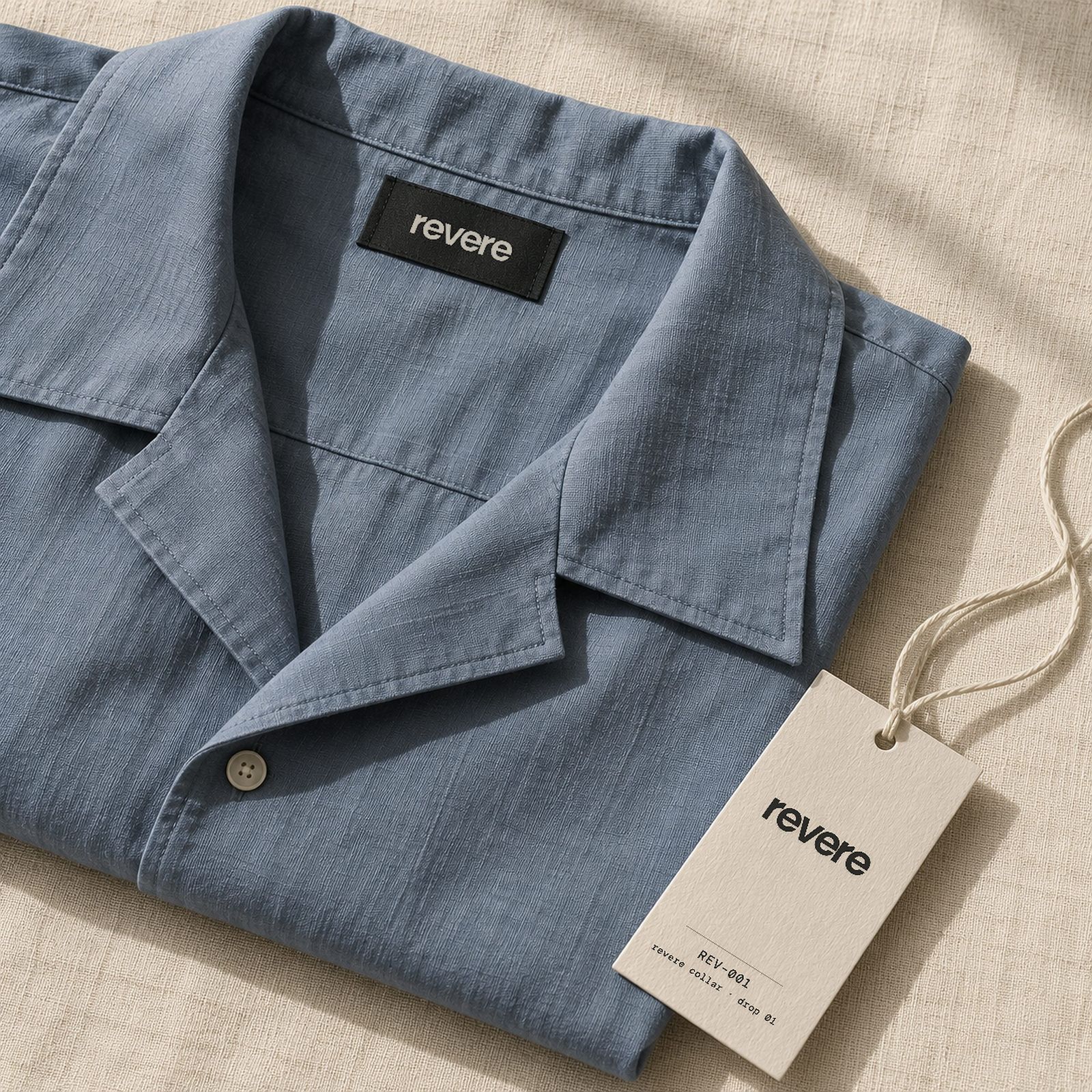 A folded slate shirt with a woven REVERE neck label and an ivory hangtag reading REV–001, revere collar, drop 01.