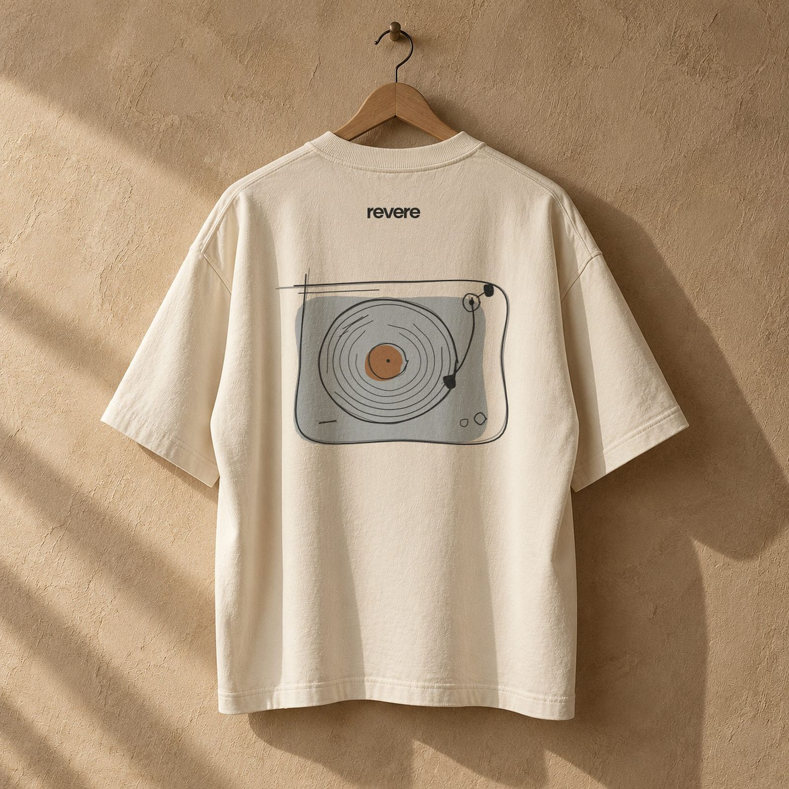 A cream heavyweight tee on a wooden hanger, printed on the back with a hand-drawn turntable and the REVERE wordmark.