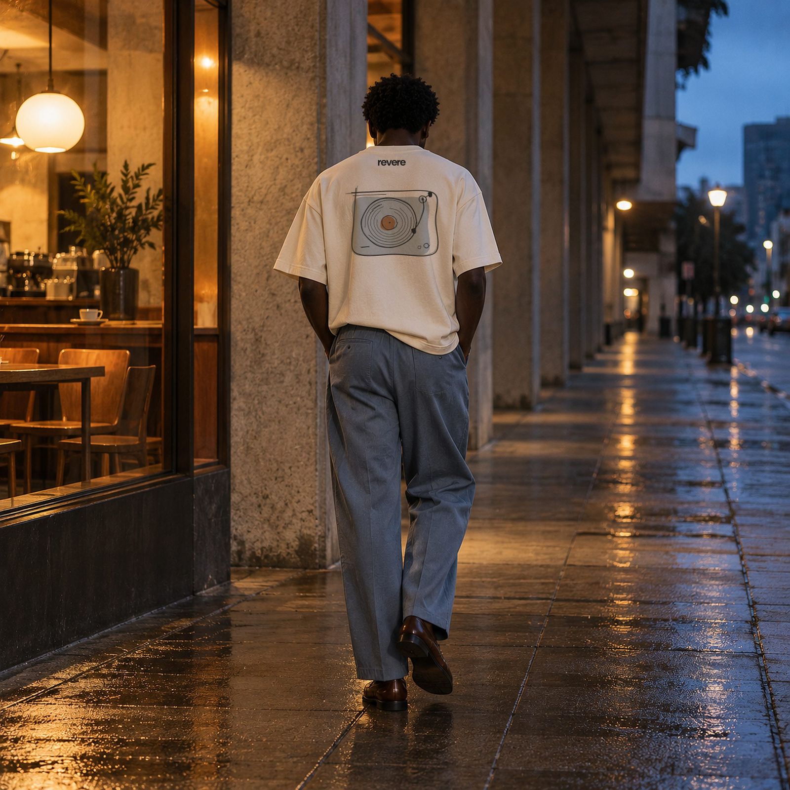 A man seen from behind at blue hour, walking past a lit café in the turntable tee and slate trousers.