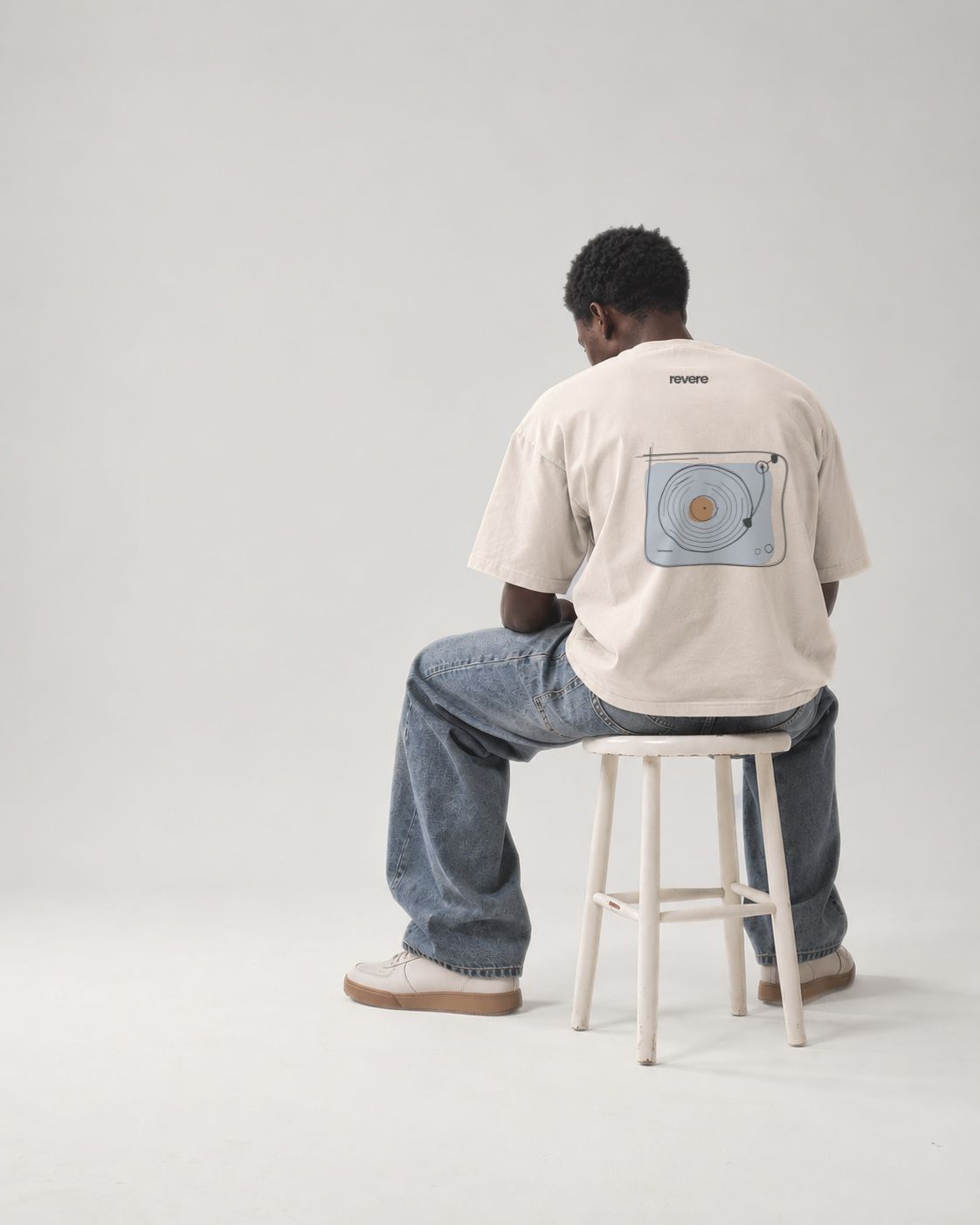 A young man seated on a white wooden stool with his back to the camera, wearing an off-white oversized T-shirt printed with a hand-drawn turntable and the REVERE wordmark, and wide blue jeans, in a pale grey studio.