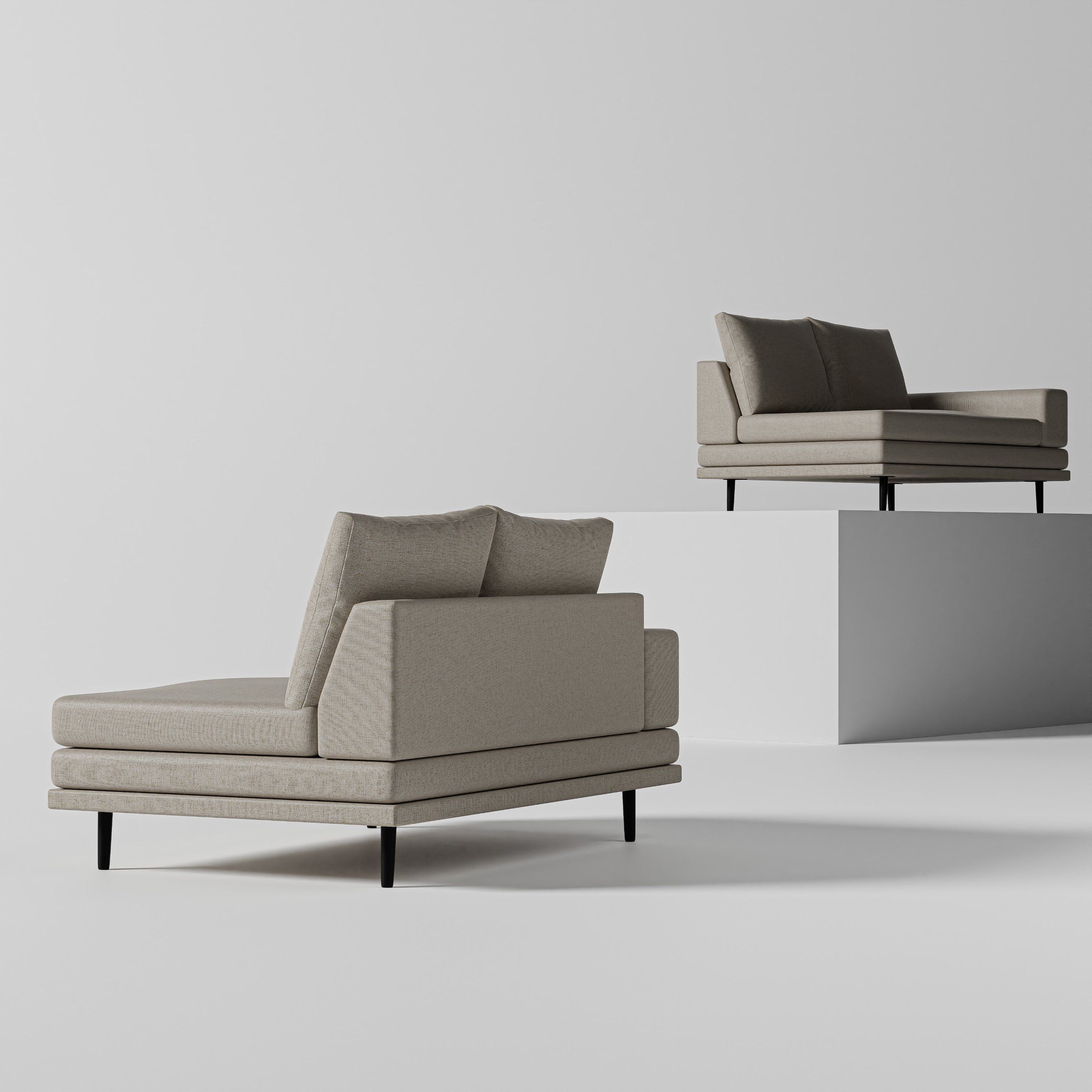 Two upholstered sofas in warm grey with thin black legs, one on the floor and one raised on a white plinth, against a pale grey backdrop.